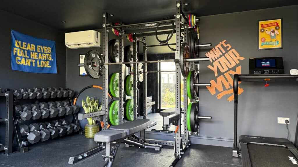 Black garden gym room fitted with a power rack, weights, bench and treadmill.