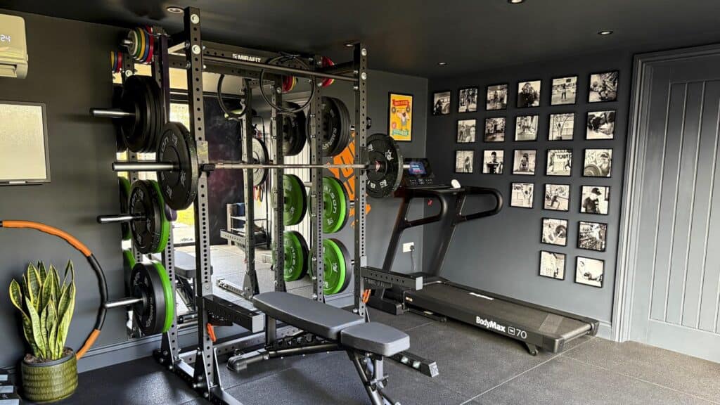 Black garden gym room fitted with a squat rack, weight plates, bench and treadmill